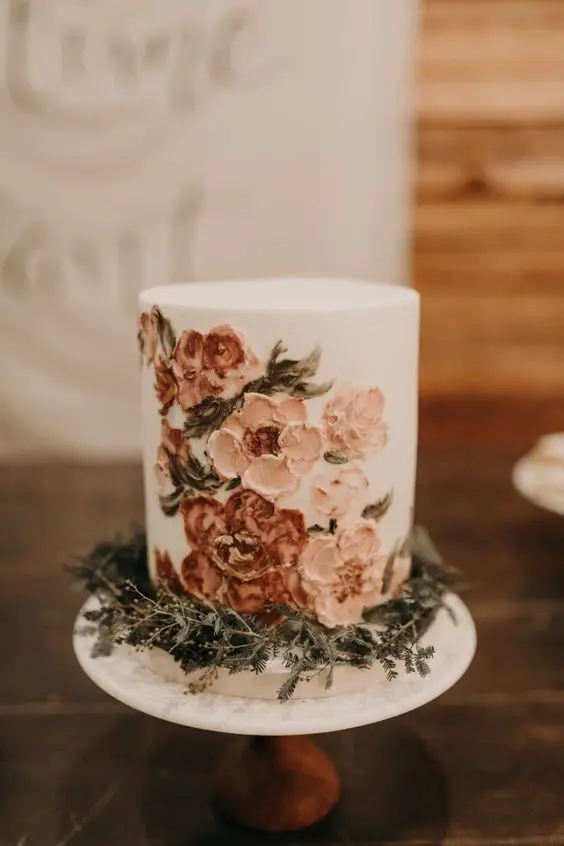 a white buttercream wedding cake with painted blush and rust blooms and greenery around the cake for a boho wedding