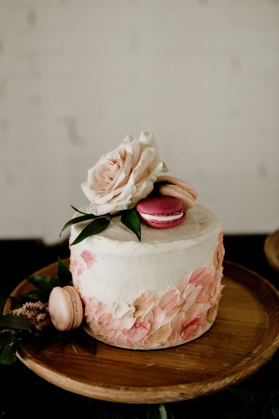 a white buttercream wedding cake with pink and blush sugar petals, blush and pink macarons on top, a blush bloom and leaves