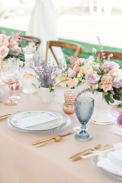 a relaxed and delicate pastel wedding tablescape with a peachy tablecloth, pink and blue glasses, pastel blooms and greenery and gold cutlery