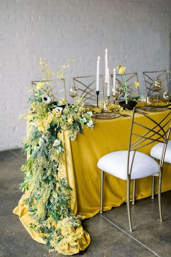 a bright and stylish spring wedding tablescape with a mustard tablecloth, bold yellow blooms and greenery and glass plates
