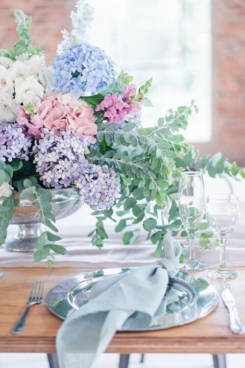 a relaxed pastel spring wedding tablescape with blue and pink hydrangeas, lilac and greenery and a light blue napkin