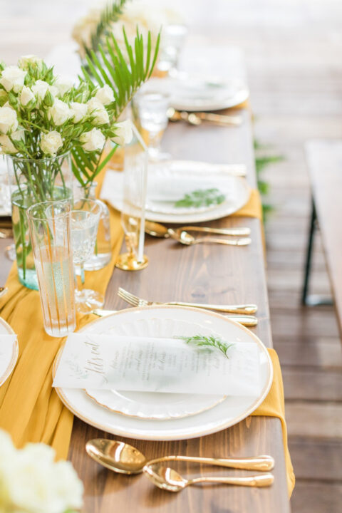 a chic and bold wedding tablescape with a yellow runner and napkins, with gold cutlery, white blooms and greenery is a vibrant idea