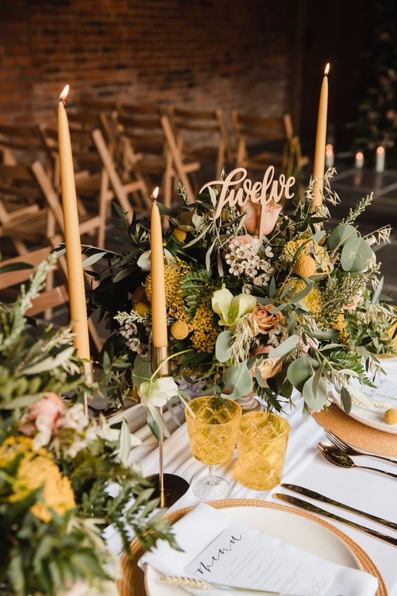 a chic wedding tablescape with yellow and neutral blooms and greenery, yellow glasses and candles, woven placemats