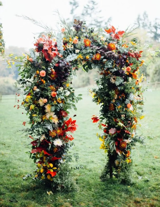 a beautiful and colorful rustic fall wedding arch with greenery, red, yellow, pink blooms, colorful fall leaves is a lovely idea for the season