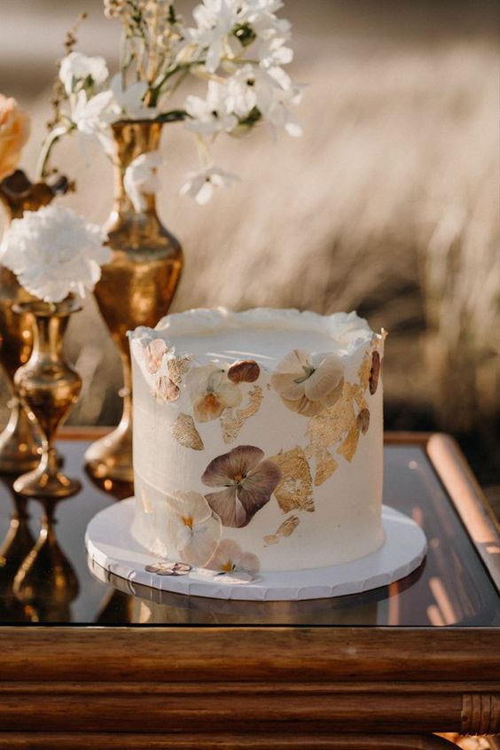 an elegant white buttercream wedding cake with gold leaf and pressed flowers is a stylish idea for a fall boho wedding