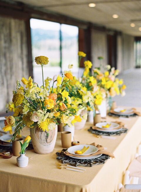 a refined wedding tablescape with yellow and rust linens, with bold yellow blooms, woven chargers and chic vases