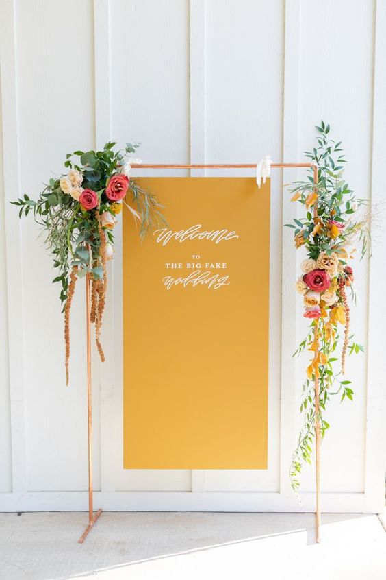 a stylish mustard wedding sign on a copper arch, with bright blooms and greenery is a stylish idea for a modern wedding