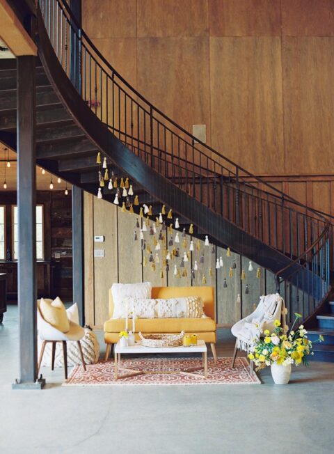 a small and cozy wedding lounge with neutral and yellow furniture, with tassles over the space, some pillows and blankets
