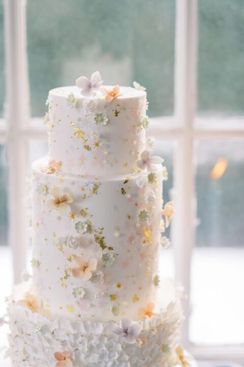 a white wedding cake decorated with tiny pastel sugar blooms, gold leaf and pearls looks very refined