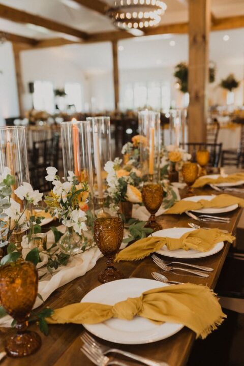 a stylish wedding tablescape with white and marigold blooms, mustard napkins, orange candles and amber glasses