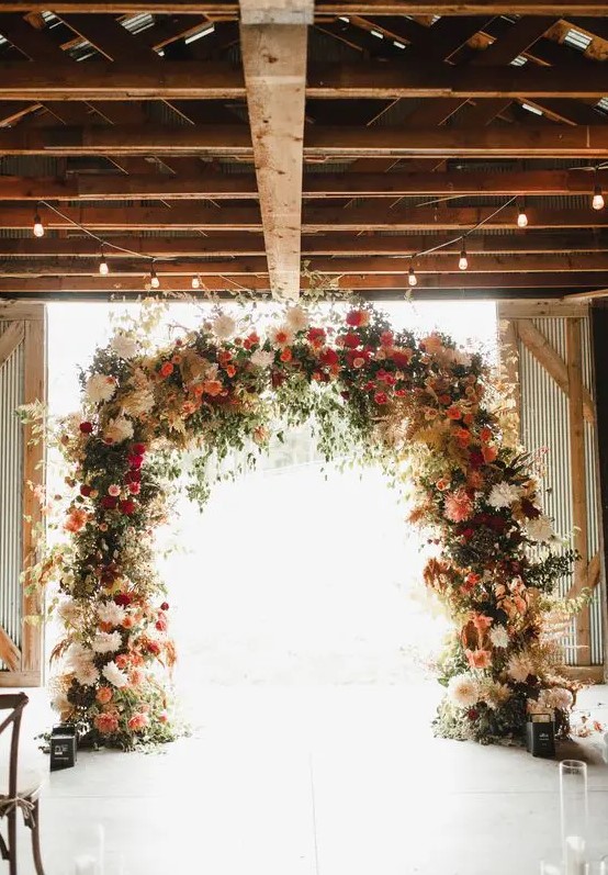 a gorgeous rustic fall wedding arch done with lots of greenery, pampas grass, blush, deep red, burgundy blooms and candle lanterns