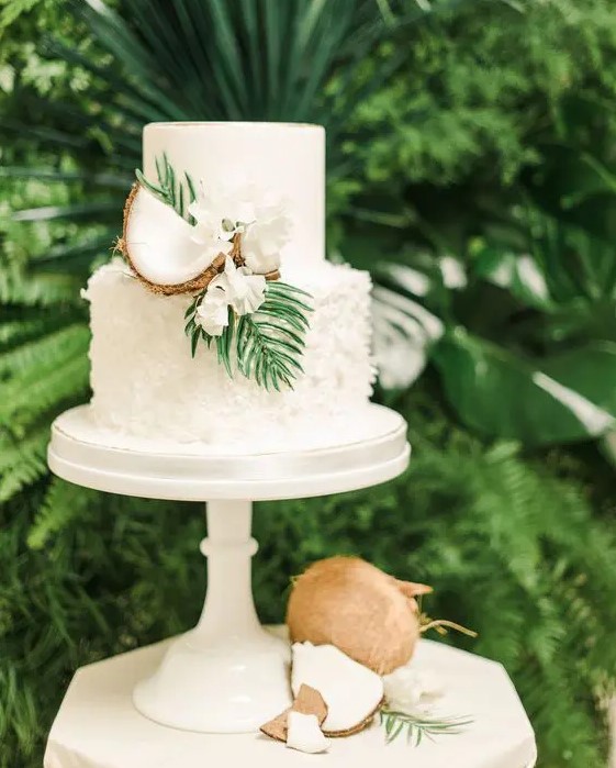 a white plain and textural wedding cake decorated with white blooms, greenery and coconuts for a tropical wedding
