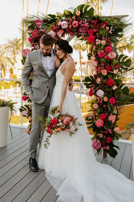 a tropical wedding arch decorated with tropical leaves, red, blush, orange and fuchsia blooms