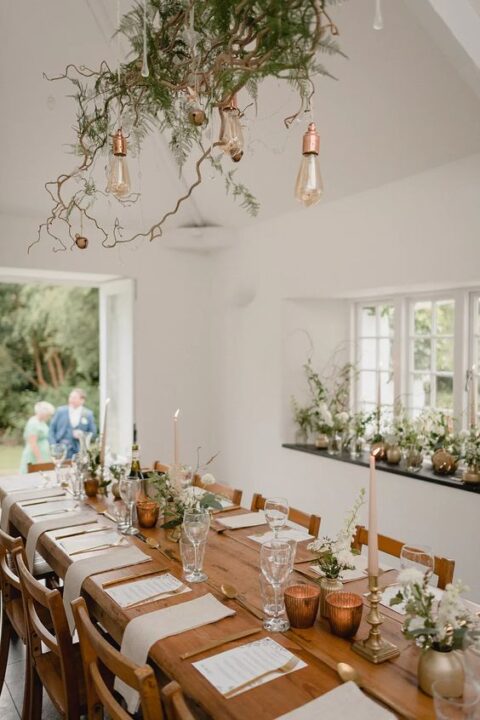 a beautiful and delicate micro wedding tablescape with white blooms and greenery, a greenery and twig arrangement over the table, blush candles and gold cutlery