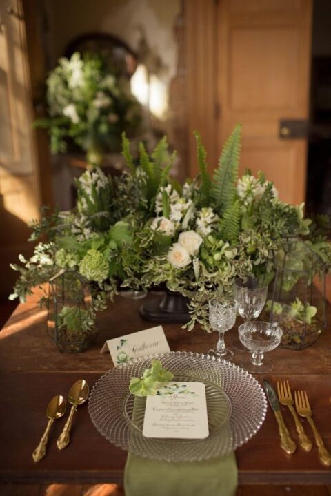 a beautiful enchanted forest wedding tablescape with neutral blooms, lots of textural greenery, green napkins, a clear charger and gold cutlery