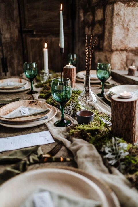 a boho enchanted forest wedding tablescape with a neutral fabric and moss runner, candles, feathers and wooden plates