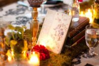 a bold enchanted forest wedding centerpiece of moss, candles, red blooms, a stack of books and a table name