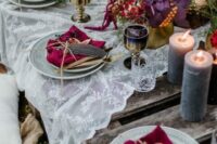a bold enchanted forest wedding table setting with a lace runner, dark and bright blooms, greenery and berries, crimson napkins, gilded glasses and lilac candles