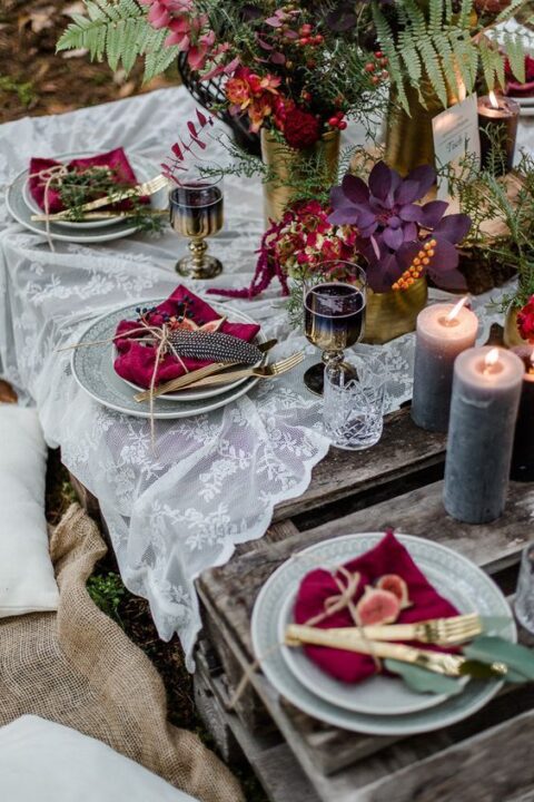 a bold enchanted forest wedding table setting with a lace runner, dark and bright blooms, greenery and berries, crimson napkins, gilded glasses and lilac candles