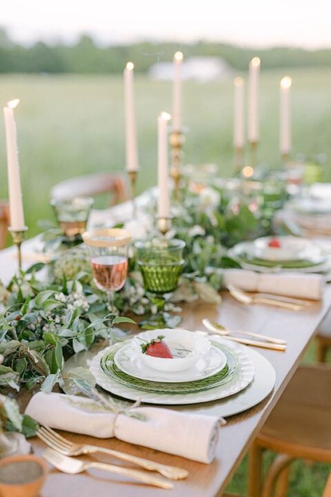 a breezy micro wedding tablescape with a lush greenery runner, green glasses and plates, blush candles and gold cutlery