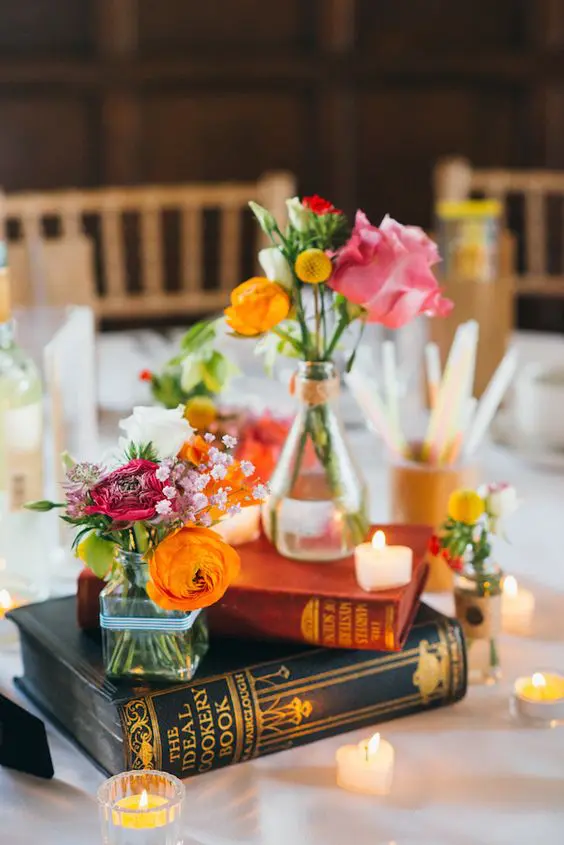 a bright eclectic wedding centerpiece of a book stack, bold blooms and greenery and some small heart shaped candles