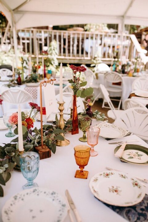 a bright enchanted forest wedding tablescape with floral print plates, a greenery runner, gilded candleholders, red and pink blooms
