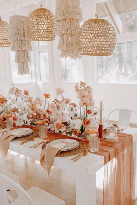 a bright micro wedding reception table with blush, white and orange blooms, an orange runner and tan napkins, woven placemats, macrame and woven lampshades