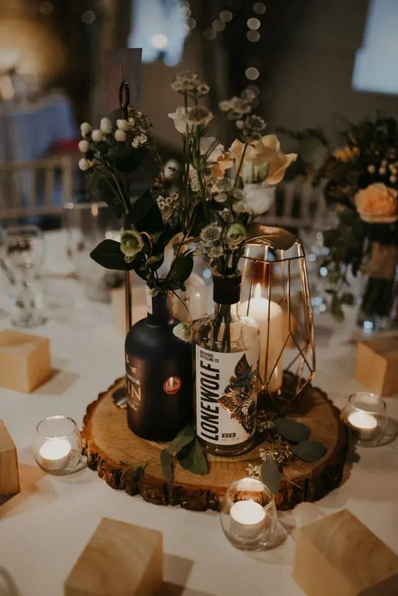 a catchy wedding centerpiece of a wood slice, bottles with greenery, berries and neutral blooms, an elegant candle lantern and candleholders around