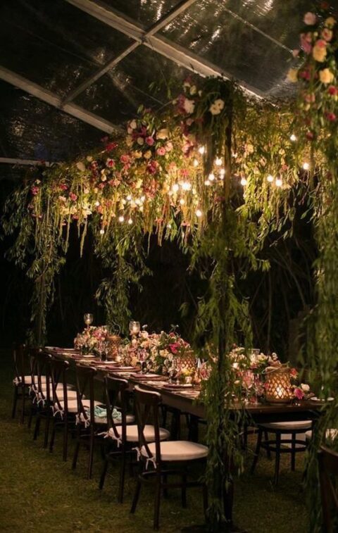 a chic indoor forest wedding reception space with lots of greenery and blooms, candles and blooms on the table and lights