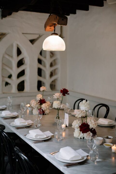 a chic modern micro wedding tablescape with white and burgundy roses, white napkins and chic plates and cutlery
