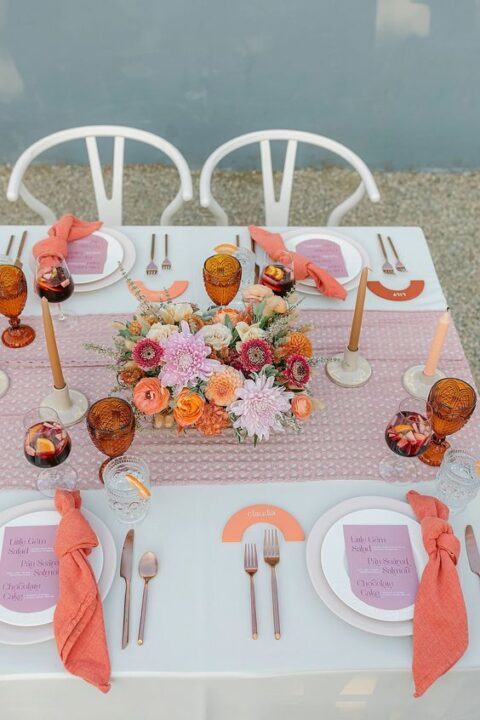 a colorful micro wedding reception table with a lilac runner, a bold floral arrangement, orange candles, coral napkins and amber glasses