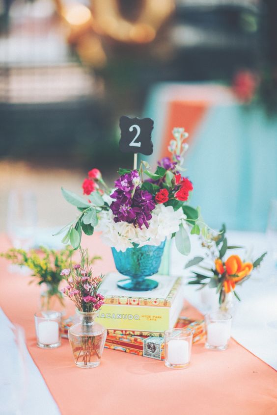 a colorful wedding centerpiece of a book stack, candleholders, small bottles with bright blooms and greenery plus a chalkboard table number