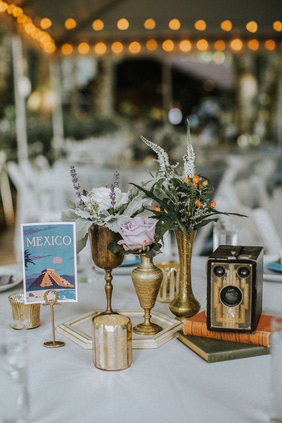 a cool travel-themed wedding centerpiece of book stacks, brass and gold vases, greenery, blooms and berries, a vintage camera and some candles