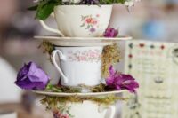 a creative wedding centerpiece of a book stack, vintage teacups with blooms, greenery and moss is a lovely idea for a wedding with a vintage feel