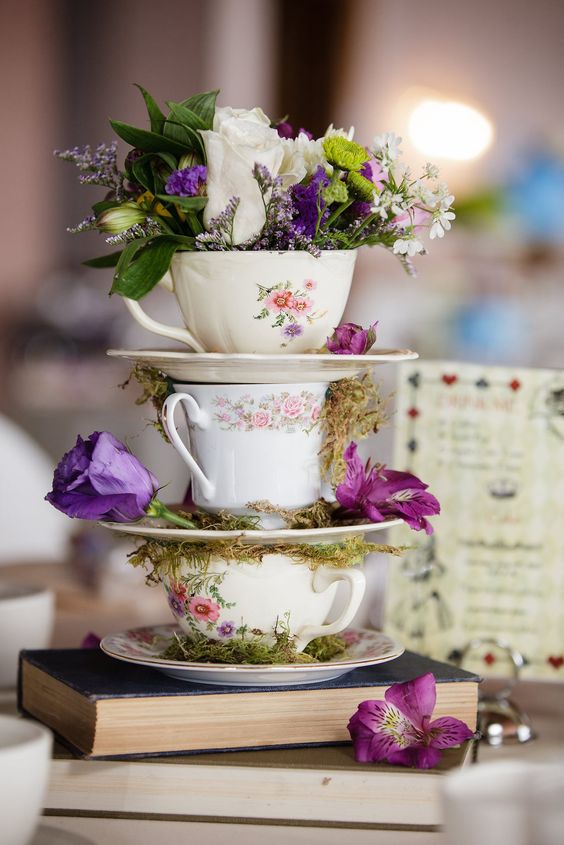 a creative wedding centerpiece of a book stack, vintage teacups with blooms, greenery and moss is a lovely idea for a wedding with a vintage feel