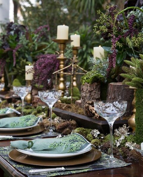 a dark enchanted forest wedding table setting done with moss, greenery, pieple blooms, pillar candles, brown placemats and green napkins, refined glasses
