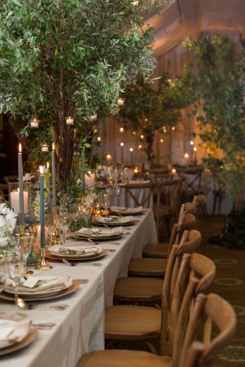 a fabulous indoor enchanted forest wedding reception space wiht trees and colored candles on the tables, some hanging candles and neutral linens
