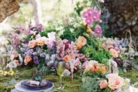a jaw-dropping enchanted forest wedding table covered with moss, bright pink, lilac and peachy blooms, lilac and purple plates and a lilac napkin, colored glasses
