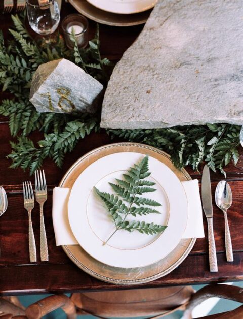a lovely enchanted forest wedding tablescape with ferns, large rocks including a gilded rock, a metallic charger and white plates