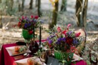 a moody enchanted forest wedding tablescape with a crimson tablecloth, super bright floral wedding centerpiece, a black runner and black candles