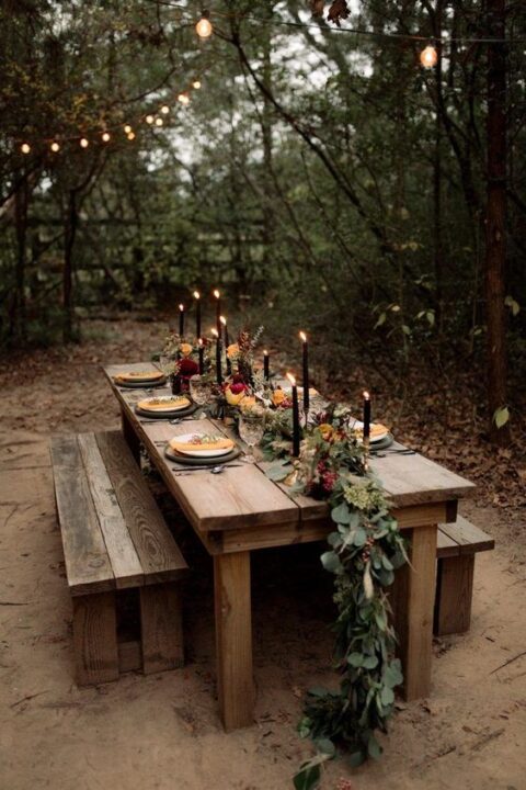 a moody rustic micro wedding table setting with a greenery and bright bloom runner, black candles, orange napkins is a chic idea