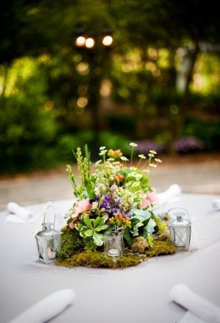 a pretty and small eclectic wedding centerpiece of moss, greenery and pastel and neutral blooms, candleholders and candle lanterns