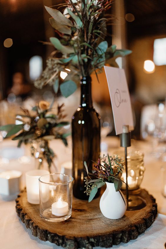 a pretty eclectic wedding centerpiece of a wood slice, candleholders, a bottle with greenery, small vases with greenery is cool