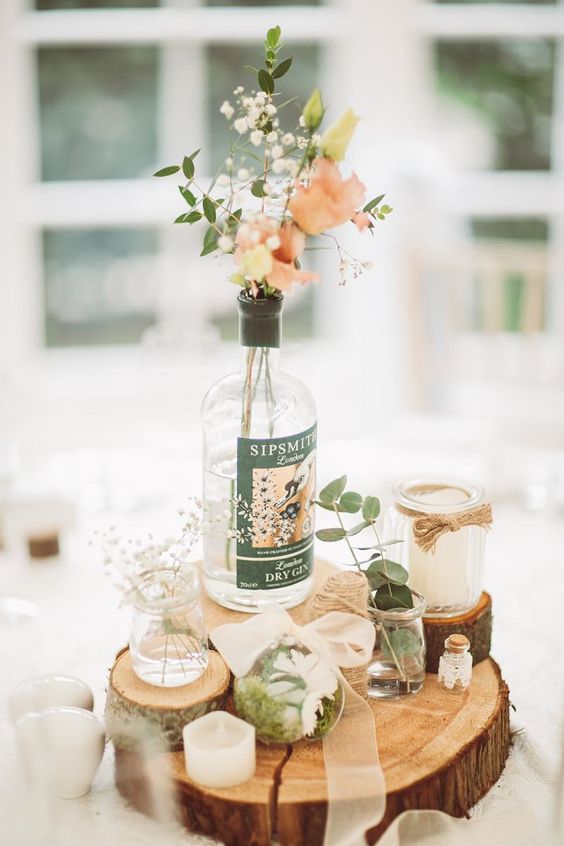 a quirky rustic wedding centerpiece of tree and branch slices, bottles and jars with pink and white blooms, greenery, moss and candles