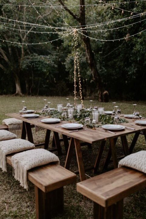 a relaxed boho micro wedding reception with an uncovered table, greenery, candles, white plates and macrame pillows on the benches