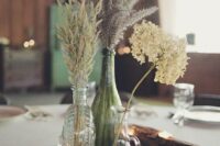 a relaxed wedding centerpiece of mismatching bottles with various grasses and blooms, a chalkboard table name, driftwood candleholders