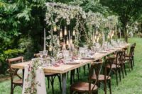 a romantic forest wedding tablescape with greenery over the table, hanging bulbs, a blush tulle and floral runner