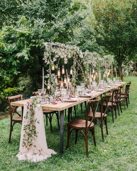 a romantic forest wedding tablescape with greenery over the table, hanging bulbs, a blush tulle and floral runner