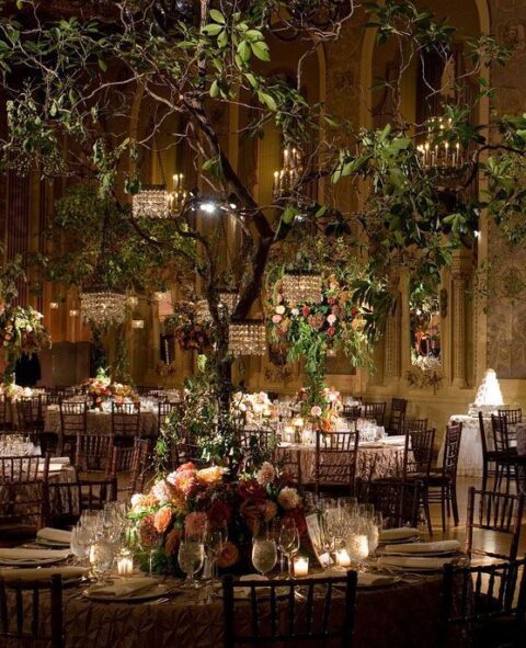 a sophisticated wedding reception space with a large tree, crystal chandeliers and blooms, lush floral centerpieces and candles