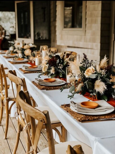 a stylish boho fall micro wedding tablescape with woven placemats, lush neutral blooms, greenery and pampas grass, candles and terracotta plates as favors
